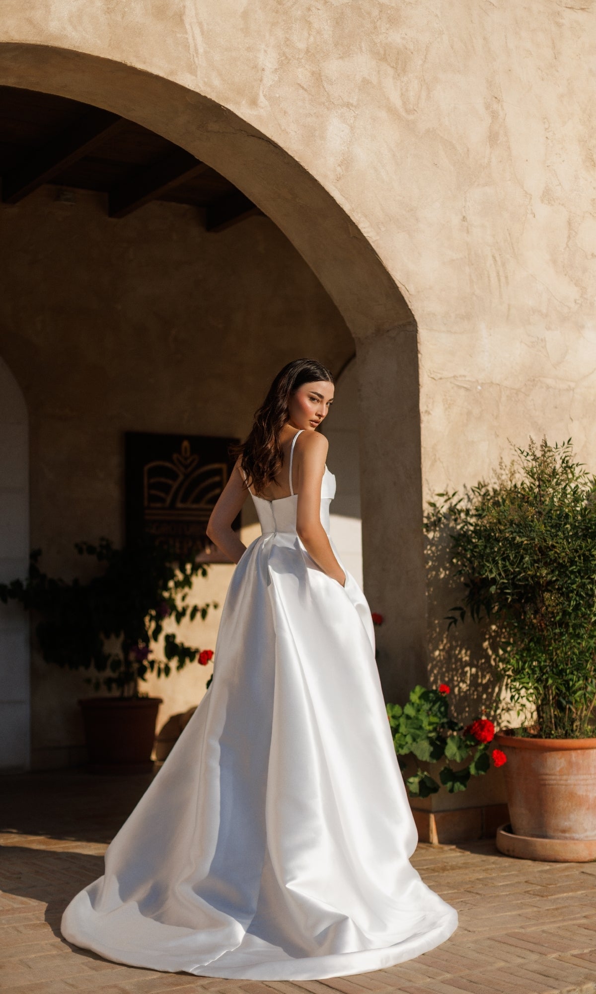 A woman in the Classic Long White Formal Dress: Tina Holly TE305 stands outdoors by an arched stone wall, looking over her shoulder.